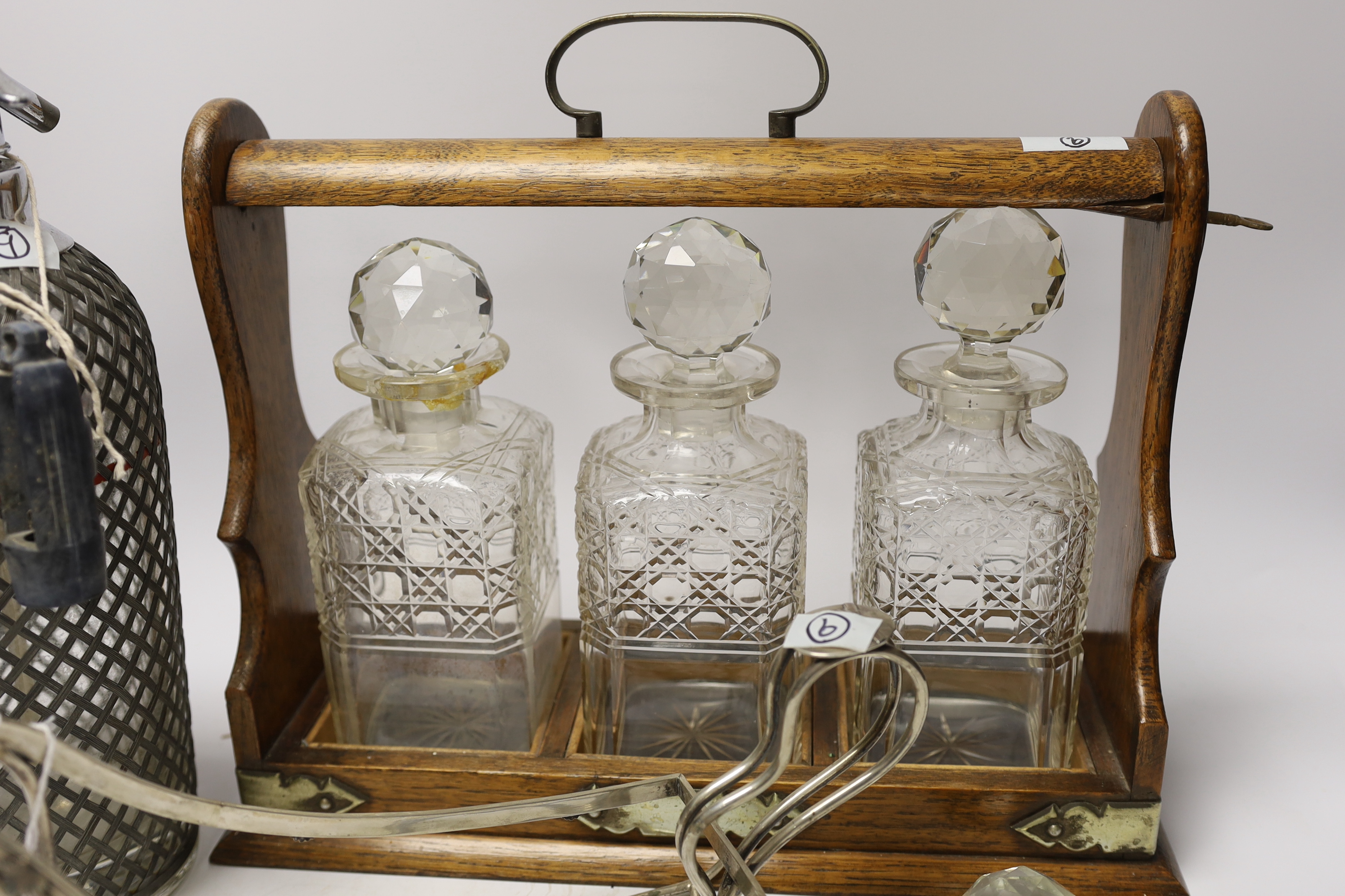 An oak three bottle tantalus, a German 800 standard white metal bottle cage and a vintage soda syphon, 36cm wide